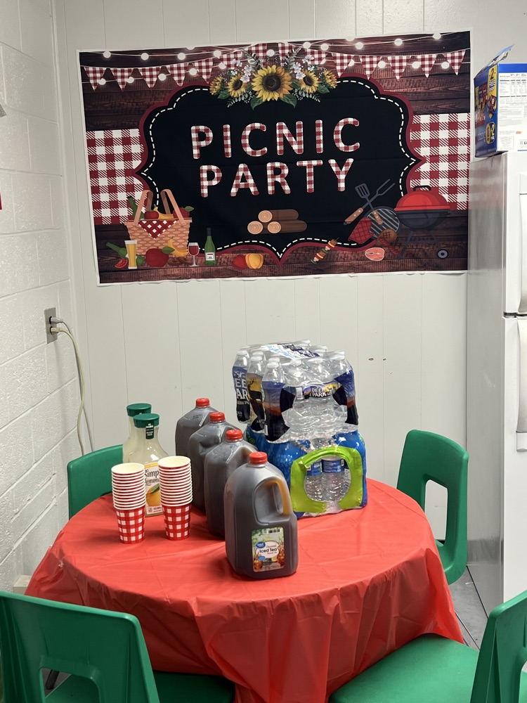 Table with red table cloth with a variety of drinks on it with a banner that says picnic party hanging in the background. 