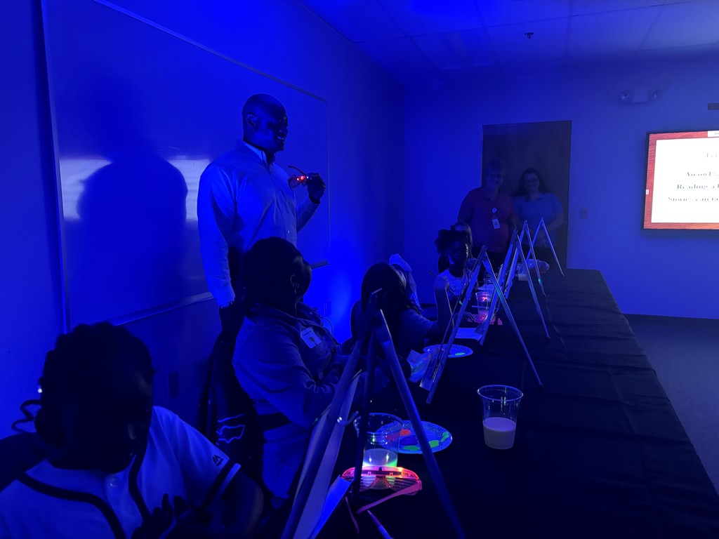 Adult holding glow in the dark glasses looks onto young artists working on paintings. 