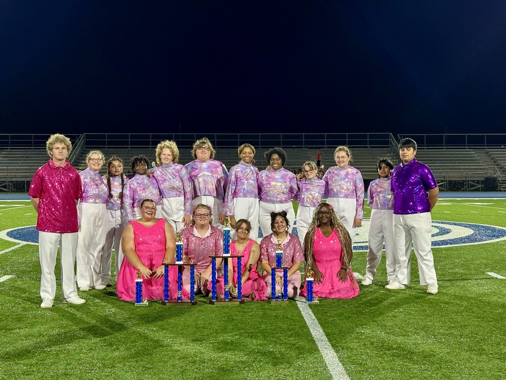 Band members in pink and purple costumes posing on field with trophies.