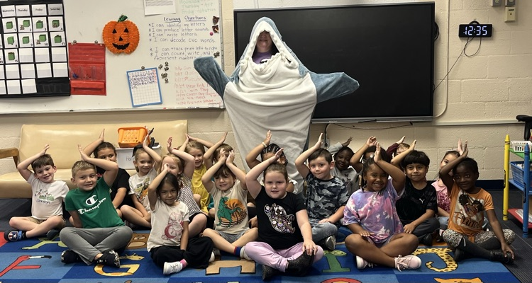 A teacher dressed up as a shark stands behind a class of students