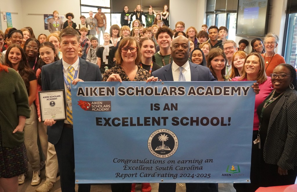 District Leadership and Aiken Scholars Academy administration and students pose with school's Excellent School Report Card Banner.