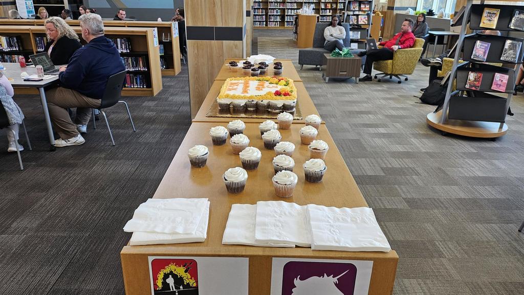 Cupcakes and Cake and napkins on a counter with people with people sitting in library. 
