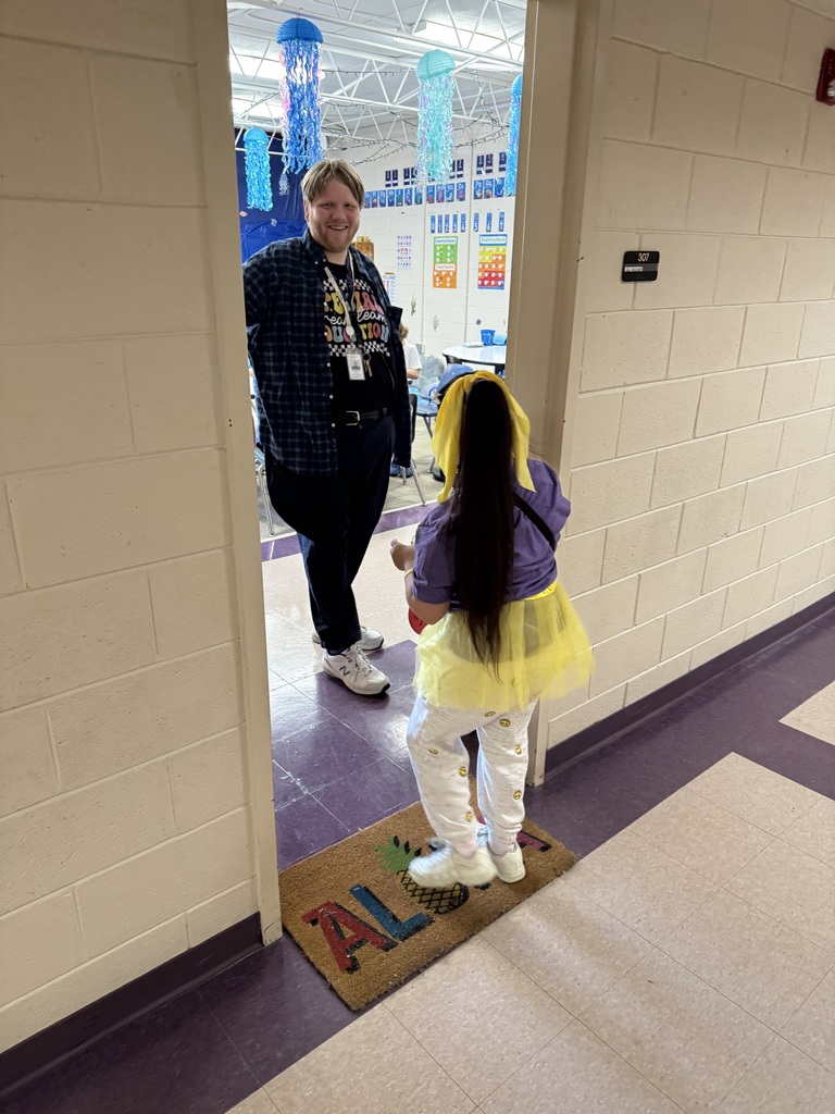 A girl wearing a yellow tutu is greeted by a teacher at a classroom door.