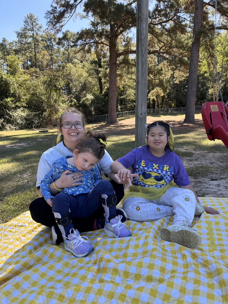 A female adult holds girl in her lap while another girl sits next to them on a blanket on the ground.