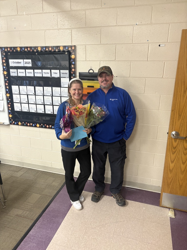 A teacher holds an award and two bouquets of flowers and stands next to her husband.