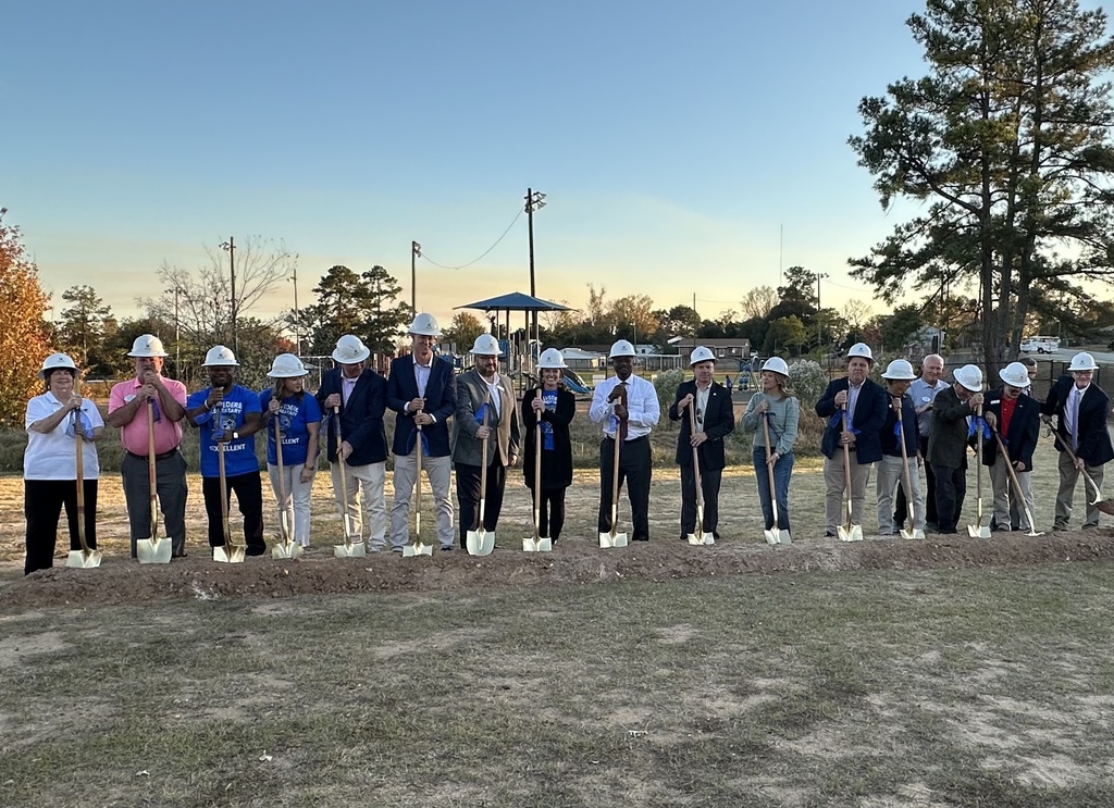 Men and Women lined up in front of shovels getting ready to break ground wearing hard hats.