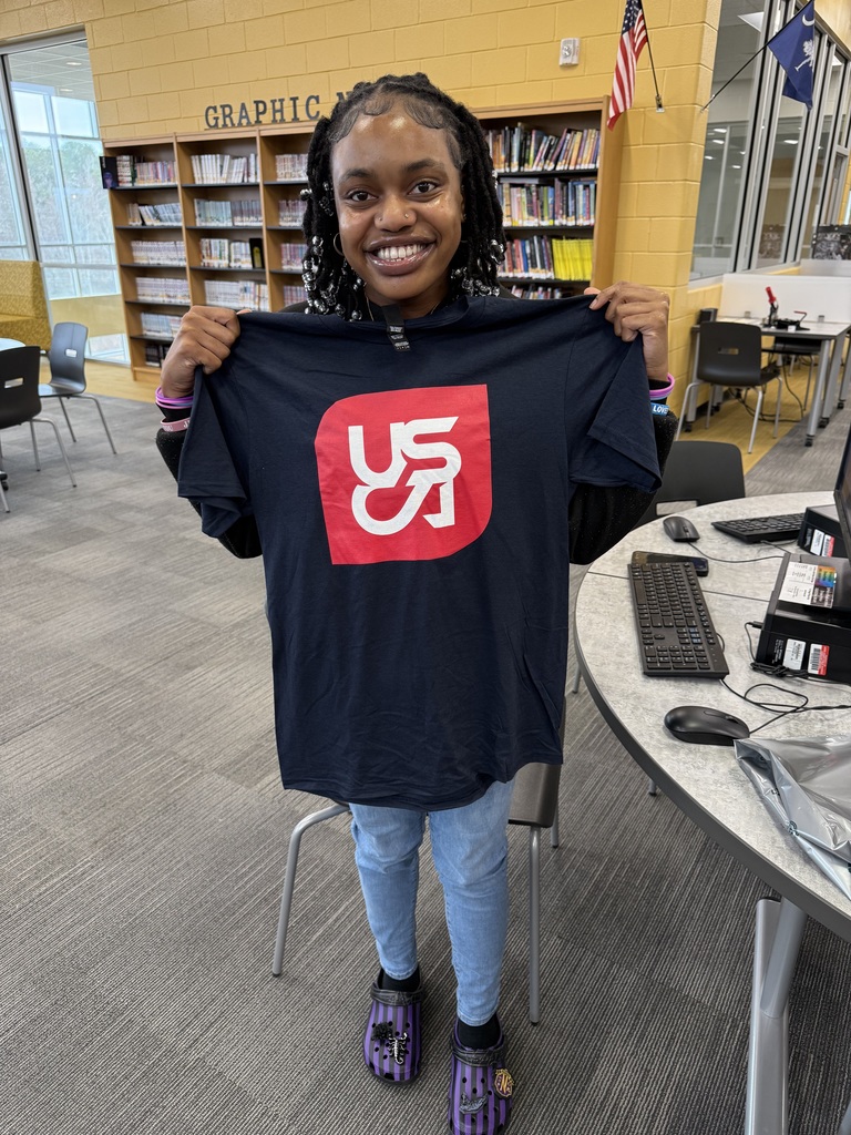 1 Student holding up USCA Shirt in library,. 