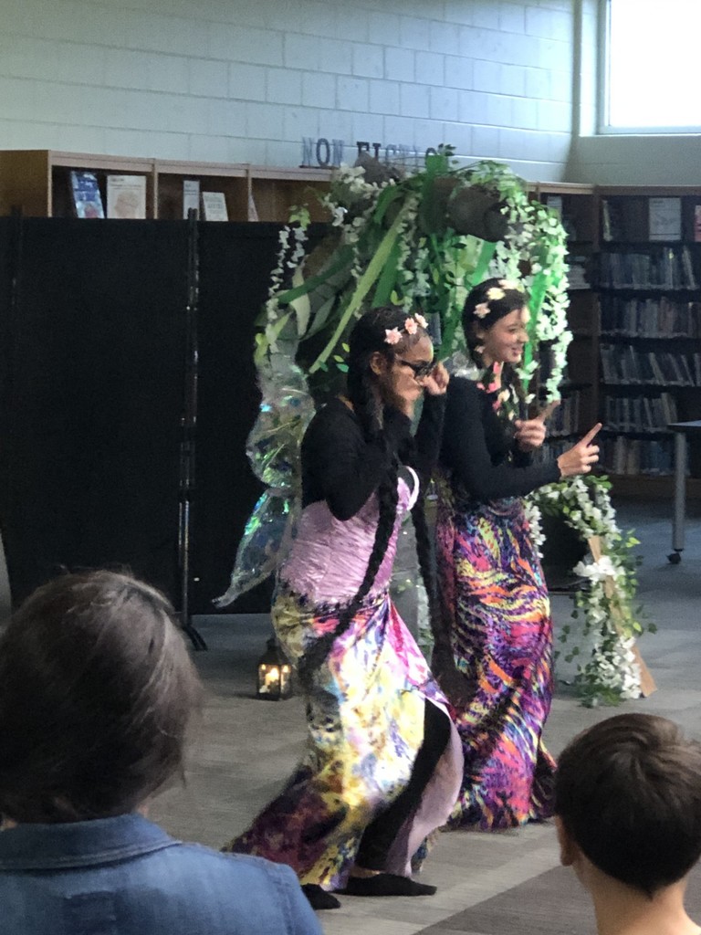 Students in costumes presenting play with sheer flowered backdrop.