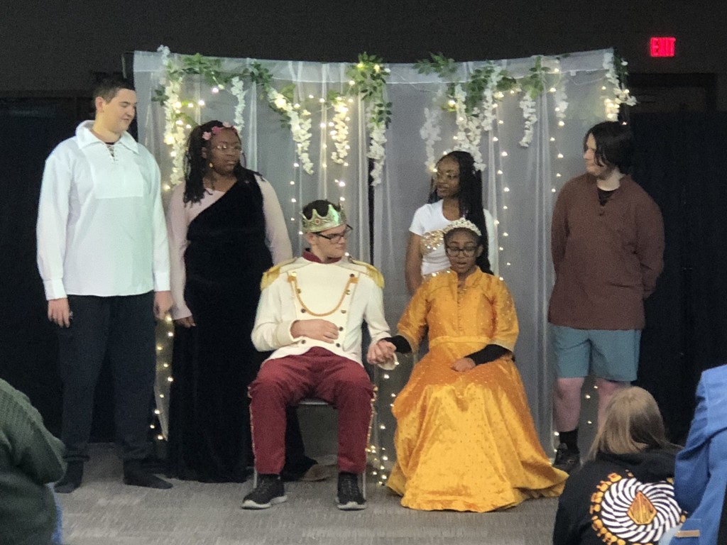 Students in costumes presenting play with sheer lighted backdrop.