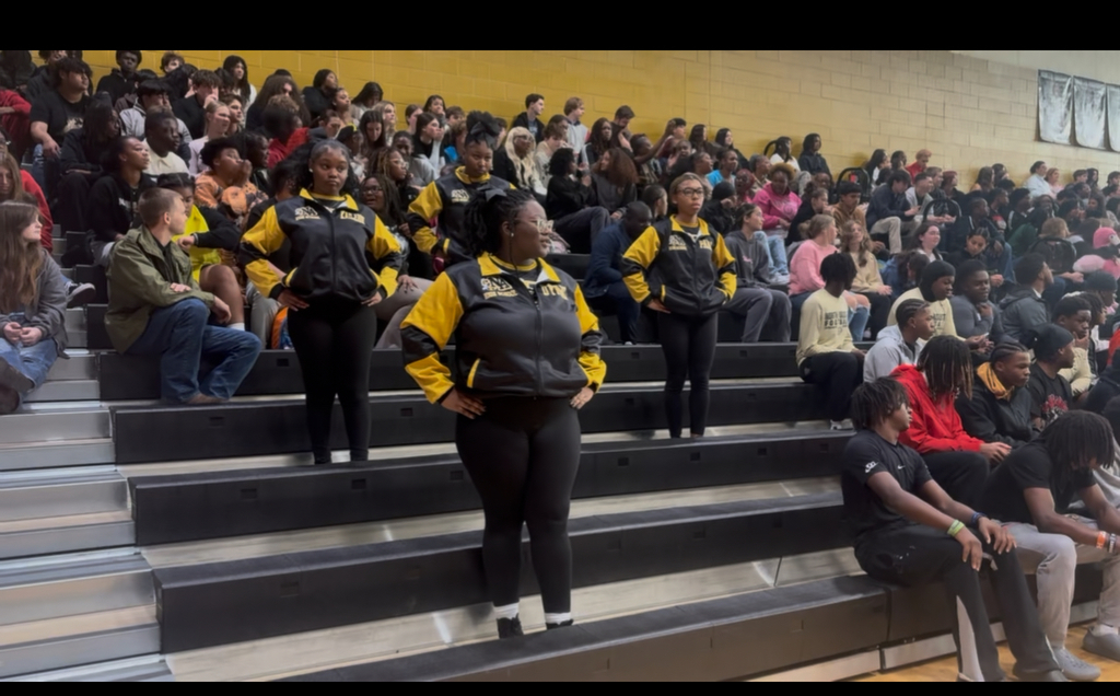 3 students dressed in black and yellow surrounded by other students sitting on bleachers.