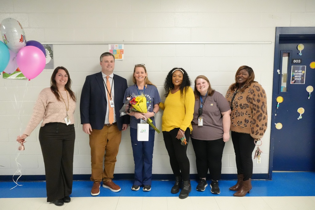 A group of six adults pose for a photo, one is holding baloons and another flowers. 
