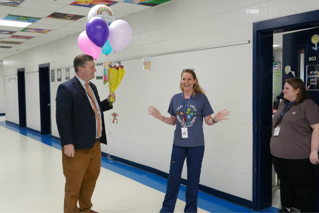 A man holds balloons and appears to be surprising a female teacher in a hallway with another person looking on. 