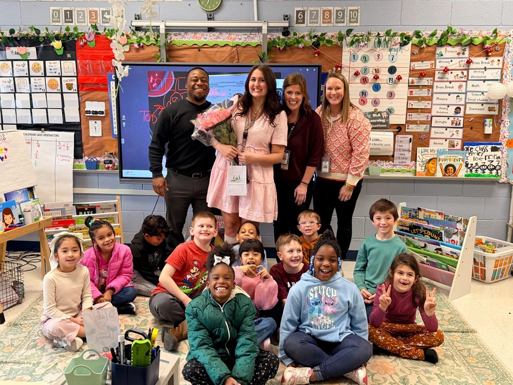A group of four adults stand behind young students in a classroom.