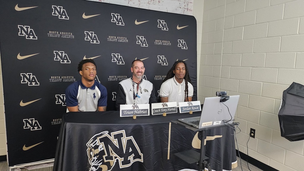 Coach and players sitting in front of display for sports in news room waiting on press conference.