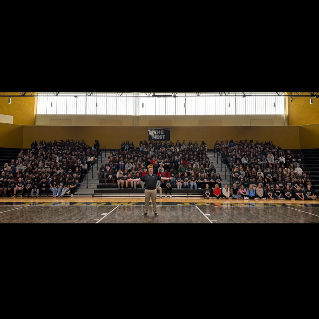 Students sitting in bleachers at gym posing with principal in front.