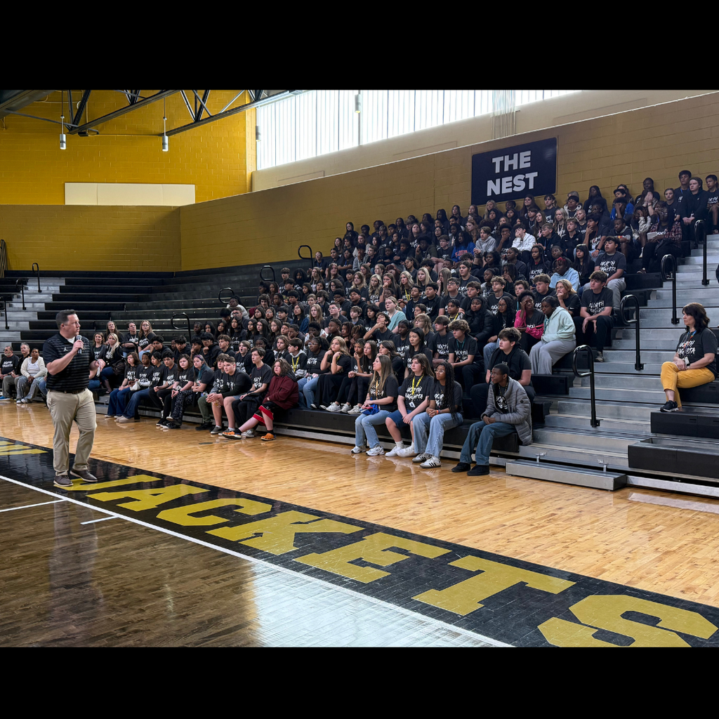 Students sitting in bleachers in gym listening to principal.