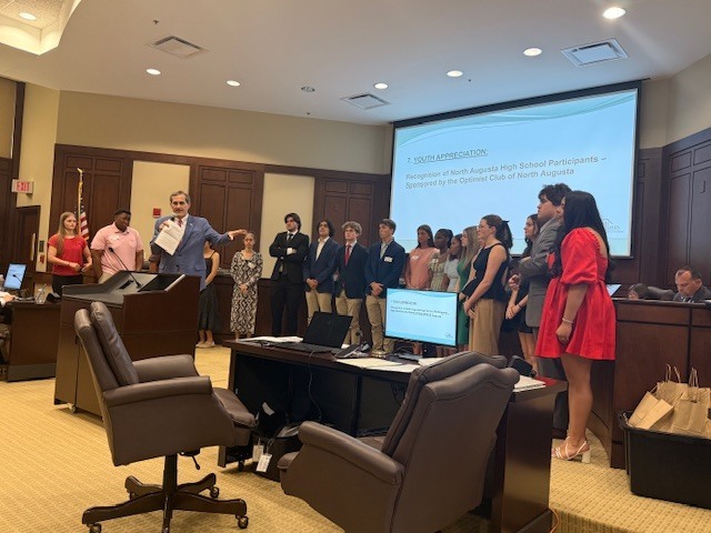Mayor speaking with students behind mayor in council chamber. 