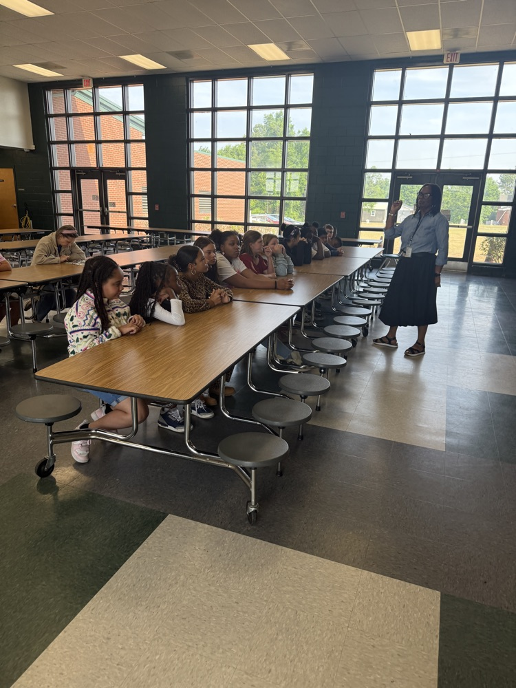 students sitting at a cafeteria table listening to an adult talk