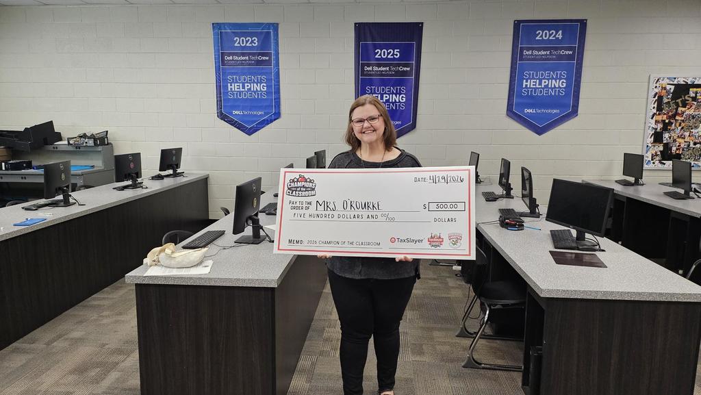 Dr. O'Rourke with Check instide computer classroom with banners in the back. 