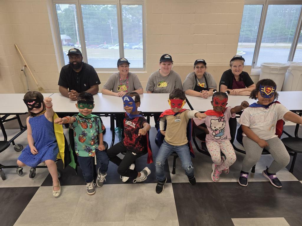 Five adult cafeteria workers sit behind six children in superhero masks and capes.