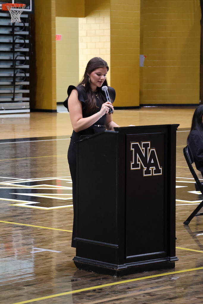 Girl standing at podium talking in gym with guests at table looking on. 