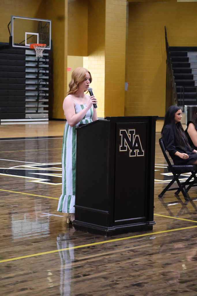 Girl standing at podium talking in gym with guests at table looking on. 