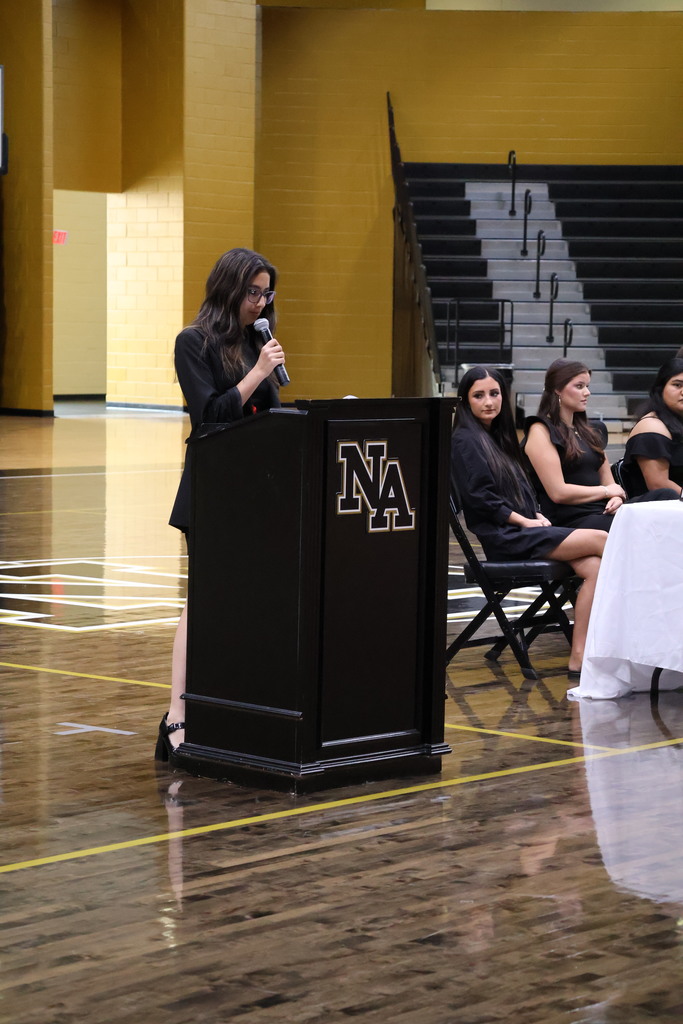 Girls standing at podium talking in gym with guests at table looking on. 