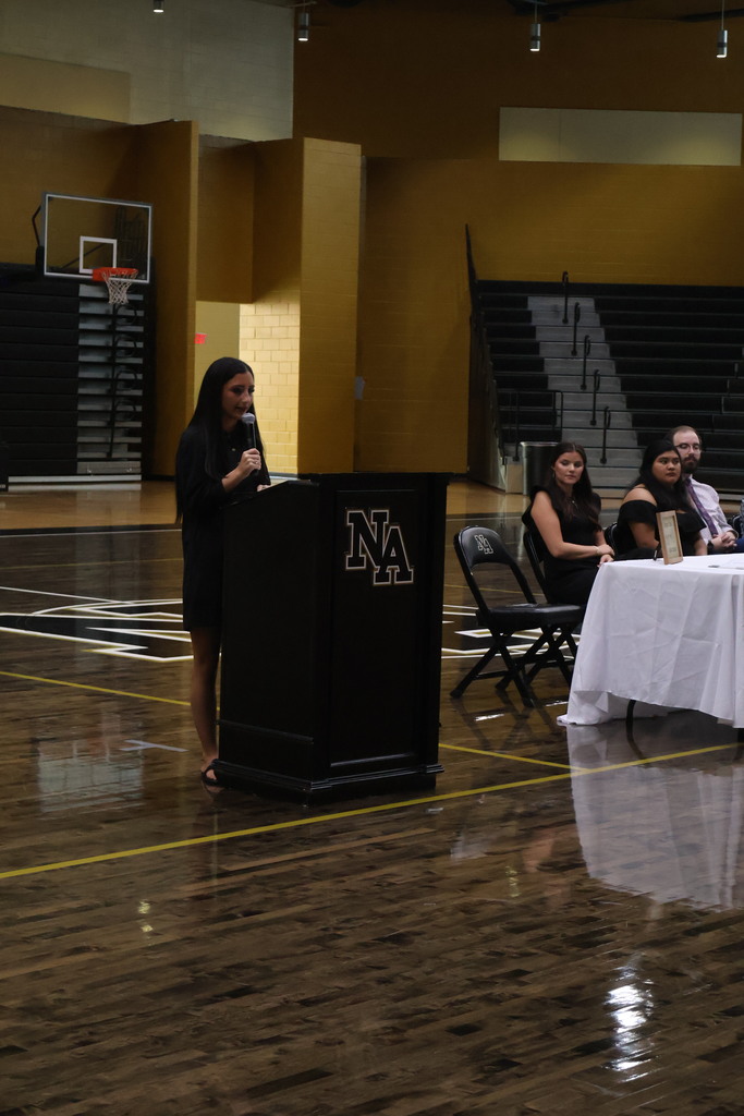 Girls standing at podium talking in gym with guests at table looking on. 