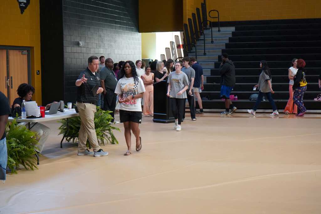 Students lined up in gym with principal with plants and tables. 