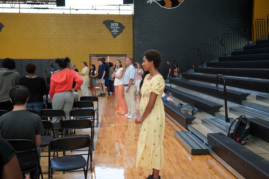 Student standing at the end of a row of students with teachers in background.