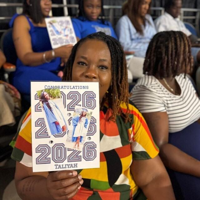 A woman is sitting holding a fan with a graduate's name and photo during graduation ceremony.