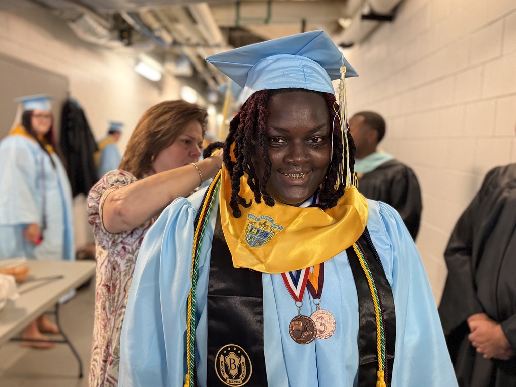 A student smiling while receiving assistance from a staff member with their graduation robe and accessories.