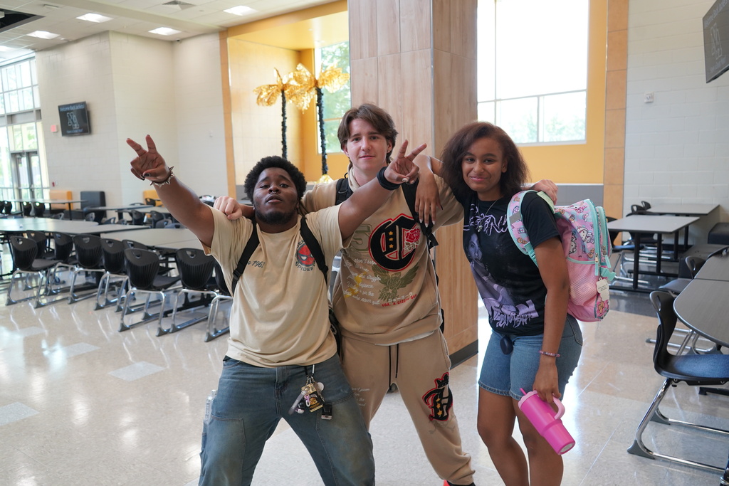 "3 students smiling for the camera giving the piece sign in a cafeteria."