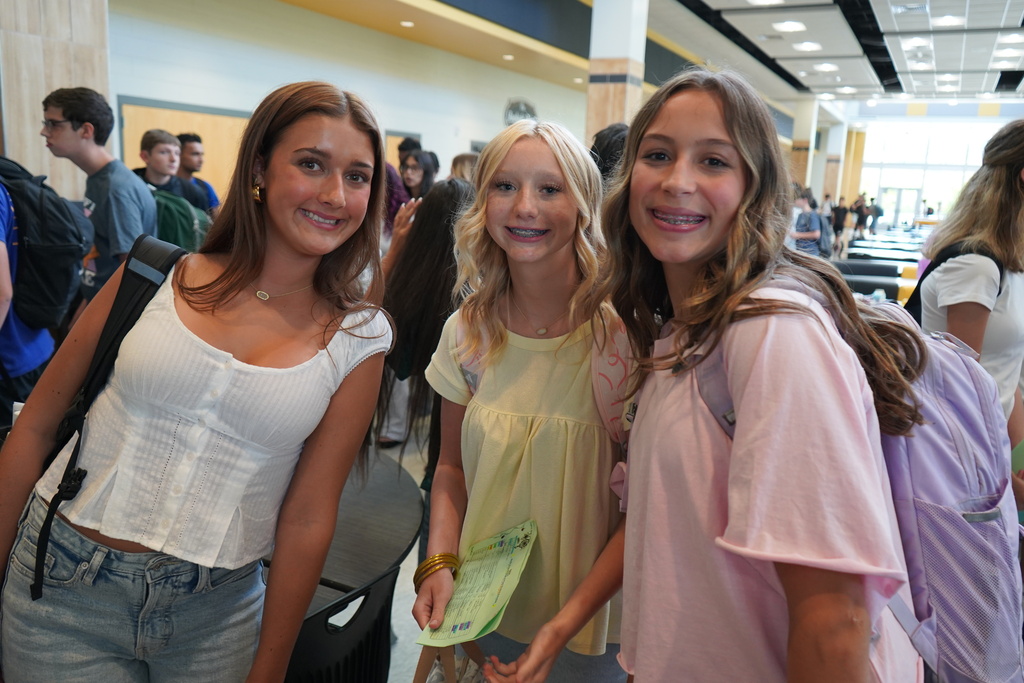 "3 girls smiling for the camera with students in the background. "