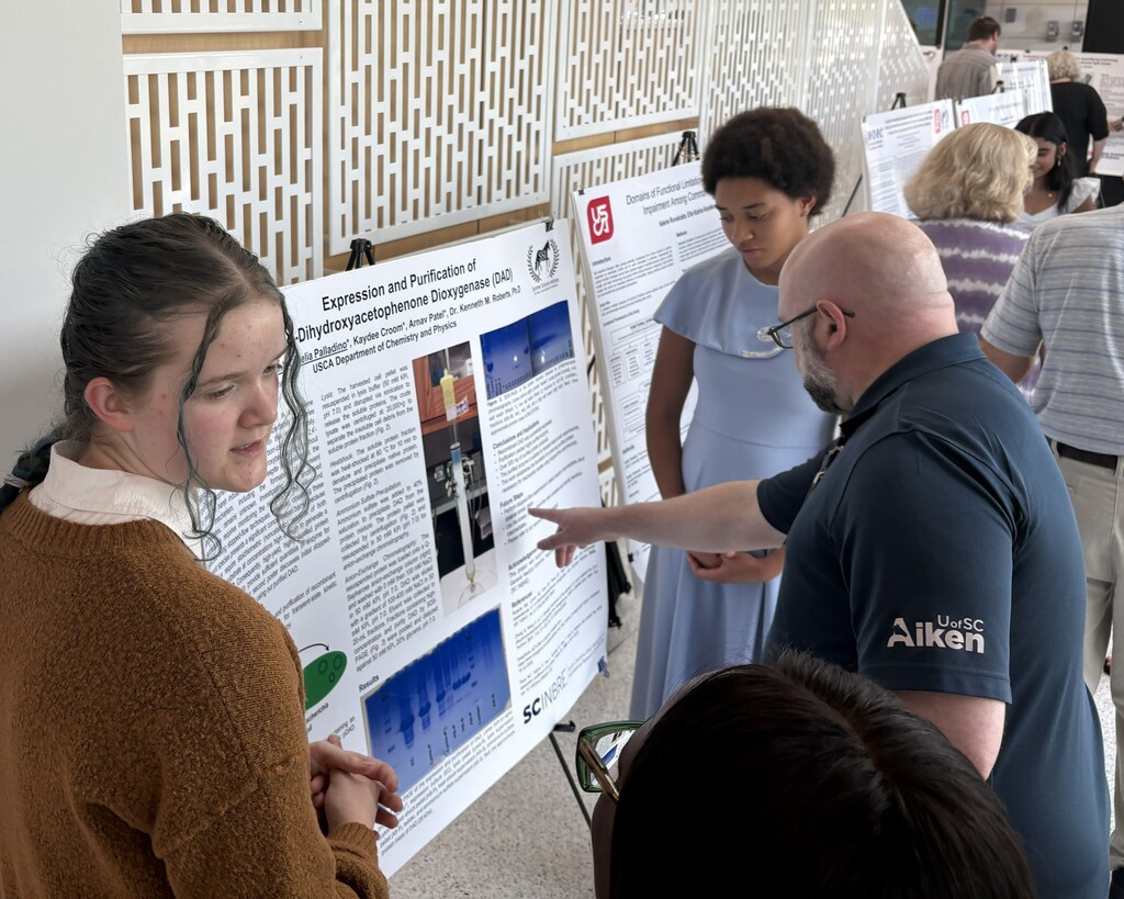 Students presenting their work on a bulletin board to a professor.  