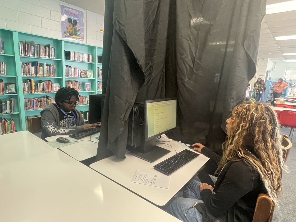 Two people using computers at a desk, with a bookshelf and a curtain in the background.