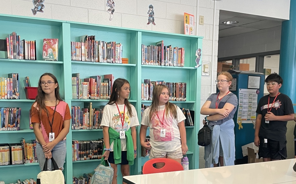 Five children stand in a school library, facing the camera. Blue shelves with books are behind them.