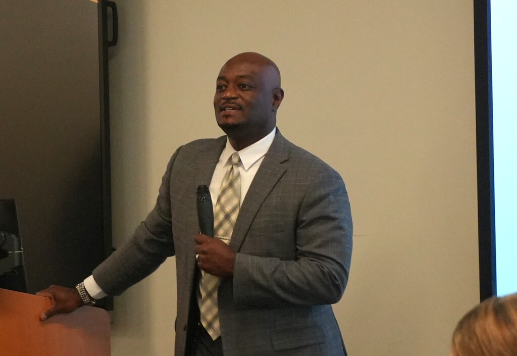 A man speaks into a microphone in a room with a projector screen in the background.