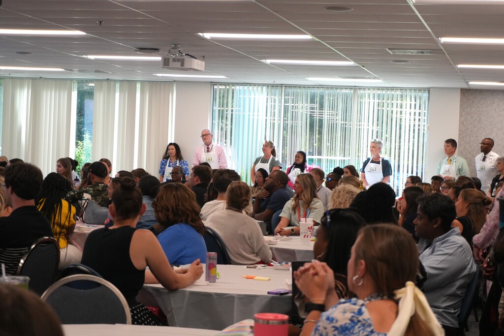 A conference room with tables and chairs. Many people sit facing the front where a few people stand.