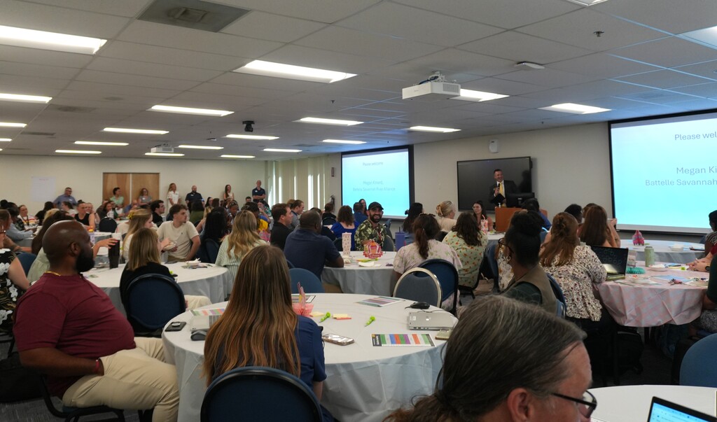 A conference room with people seated at round tables. A person is speaking in front of a screen.