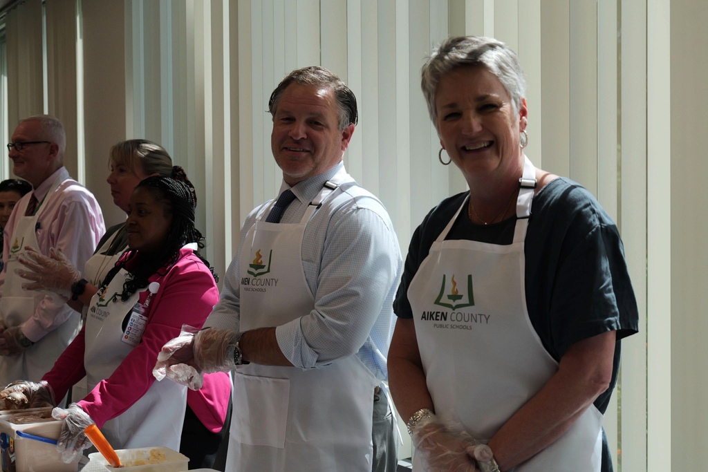 A man and woman wear aprons and gloves, smiling during the Induction Academy Ice Cream social. Other people in aprons and gloves are behind them.
