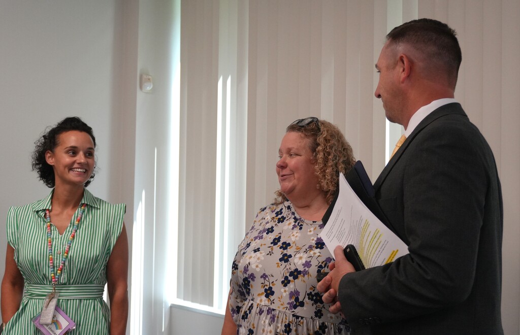 Three people in a bright room with white walls and windows. Two women and a man, one holding a folder. One of these individuals is 2026-2027 First Year Teacher of the Year Aleesha Ashe.