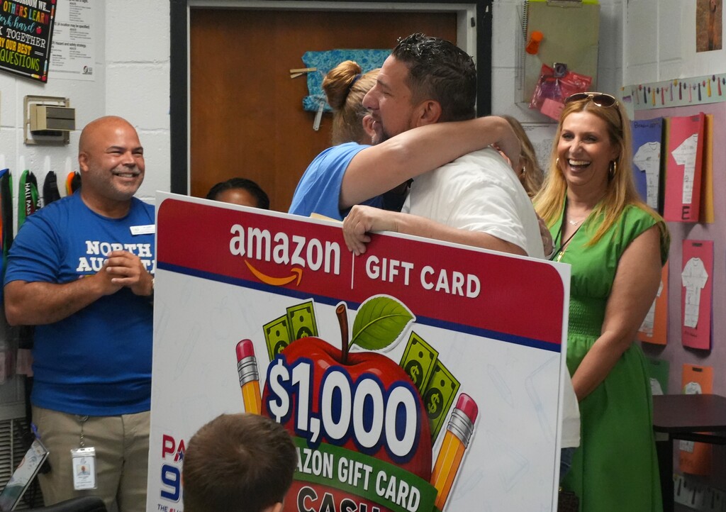 A man in a blue shirt and a woman in green hug a man in white. A large sign displays a $1,000 Amazon Gift Card.