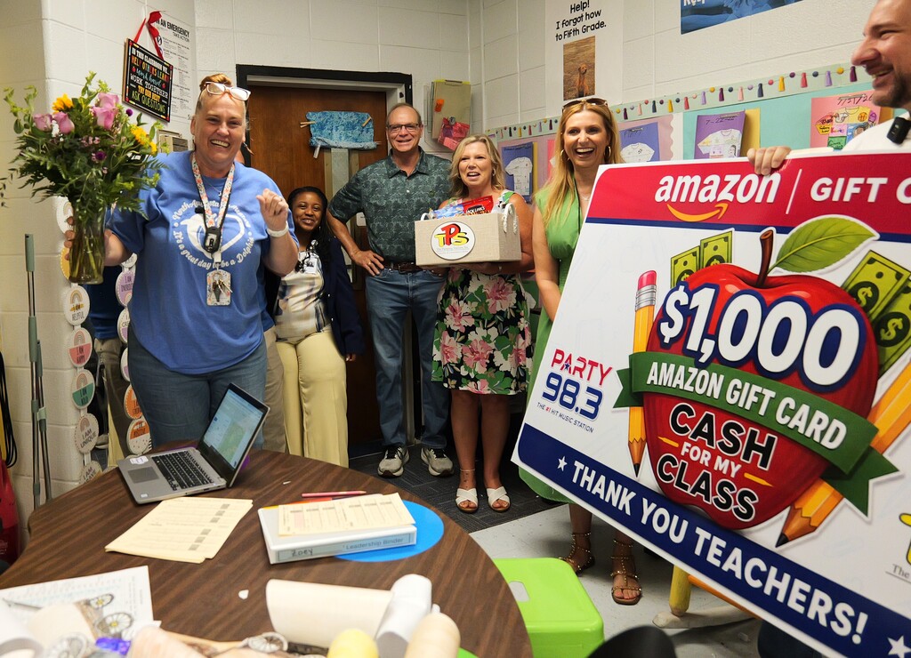 A group of adults and children in a classroom celebrate. A woman holds flowers. A man presents a box.