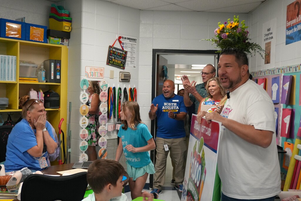 A classroom with a man holding a sign, a flower arrangement on his head, and several people looking on.
