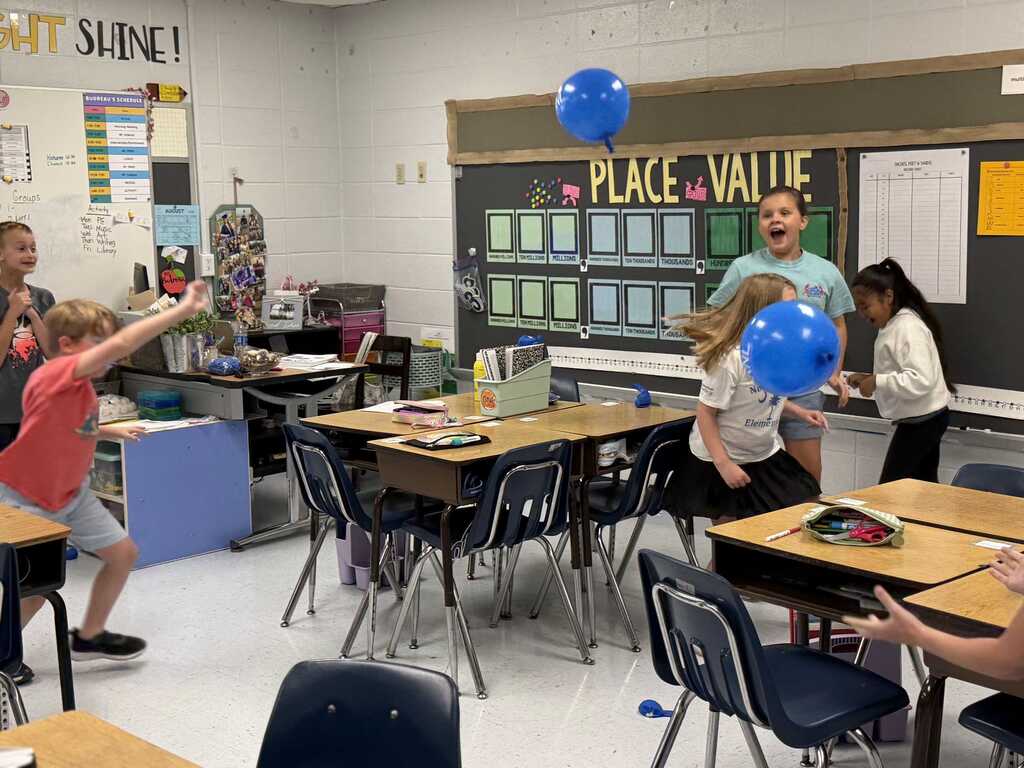 Children play in a classroom with desks, chairs, and a blackboard. Some hold balloons, and others stand near the board.
