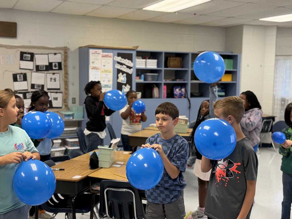 Kids holding blue balloons in a classroom with white walls, ceiling lights, and tables with chairs.
