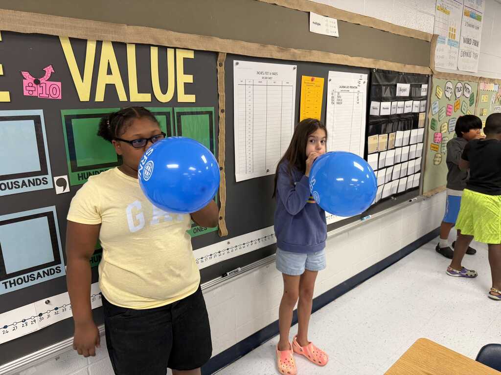 Two students blowing up blue balloons in front of a bulletin board that says "VALUE".