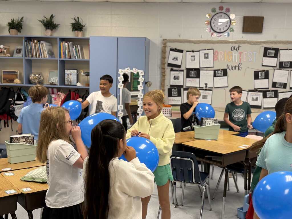 Several children in a classroom with blue balloons, standing in a circle, some holding balloons. A clock, bulletin board, and shelves with books are in the background.