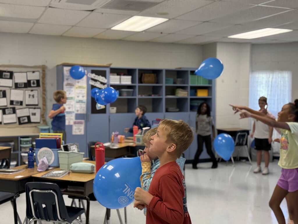 Children in a classroom play with blue balloons while a few adults observe. Desks, chairs, and shelves are in the background.